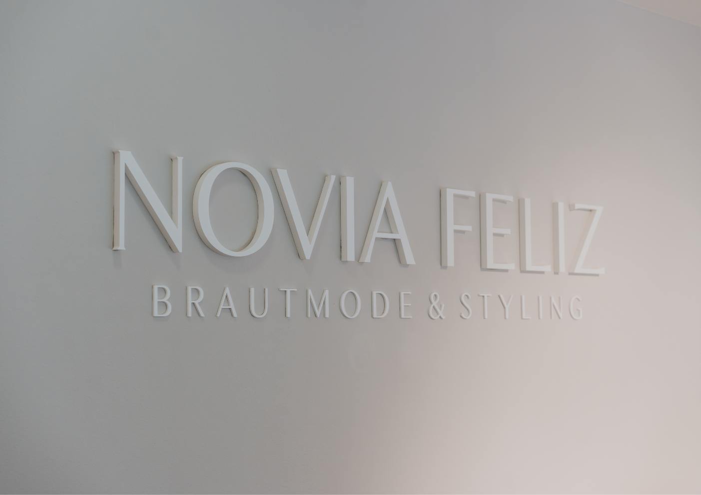 logo von novia feliz brautmode & styling in weißen buchstaben auf hellgrauer wand